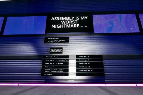 Image from the game Satisfactory featuring an industrial building surrounded by a tall security fence made from conveyor belts filled with resources from the game. This shot features a series of signs making a joke on the topic of "assembly" being both the name of a low-level programming language and a reference to one of the central gameplay mechanics of factory automation games such as Satisfactory.